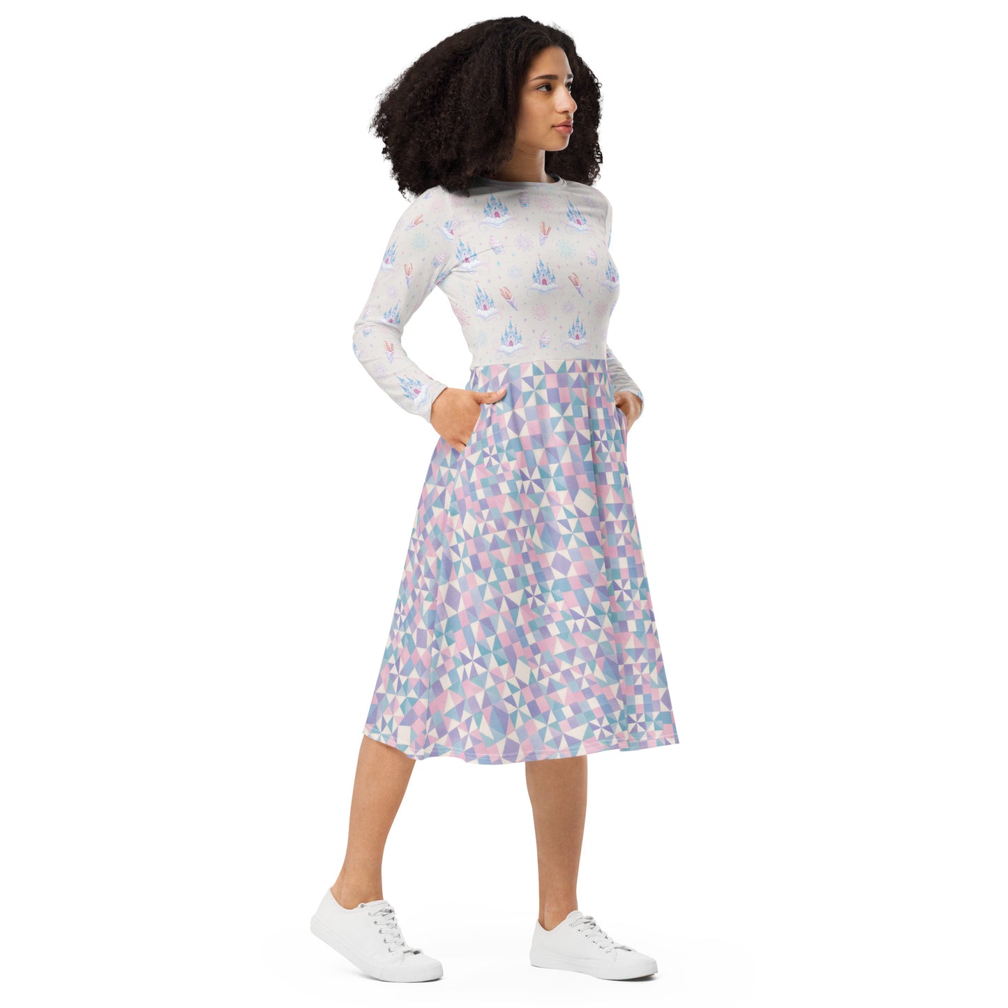 A woman wearing a long-sleeve midi dress with castles, churros, fireworks and ice cream scattered across the top and a geometric skirt pattern in pastel pink, blue, and purple on a white base with her hands tucked into the pockets of the skirt
