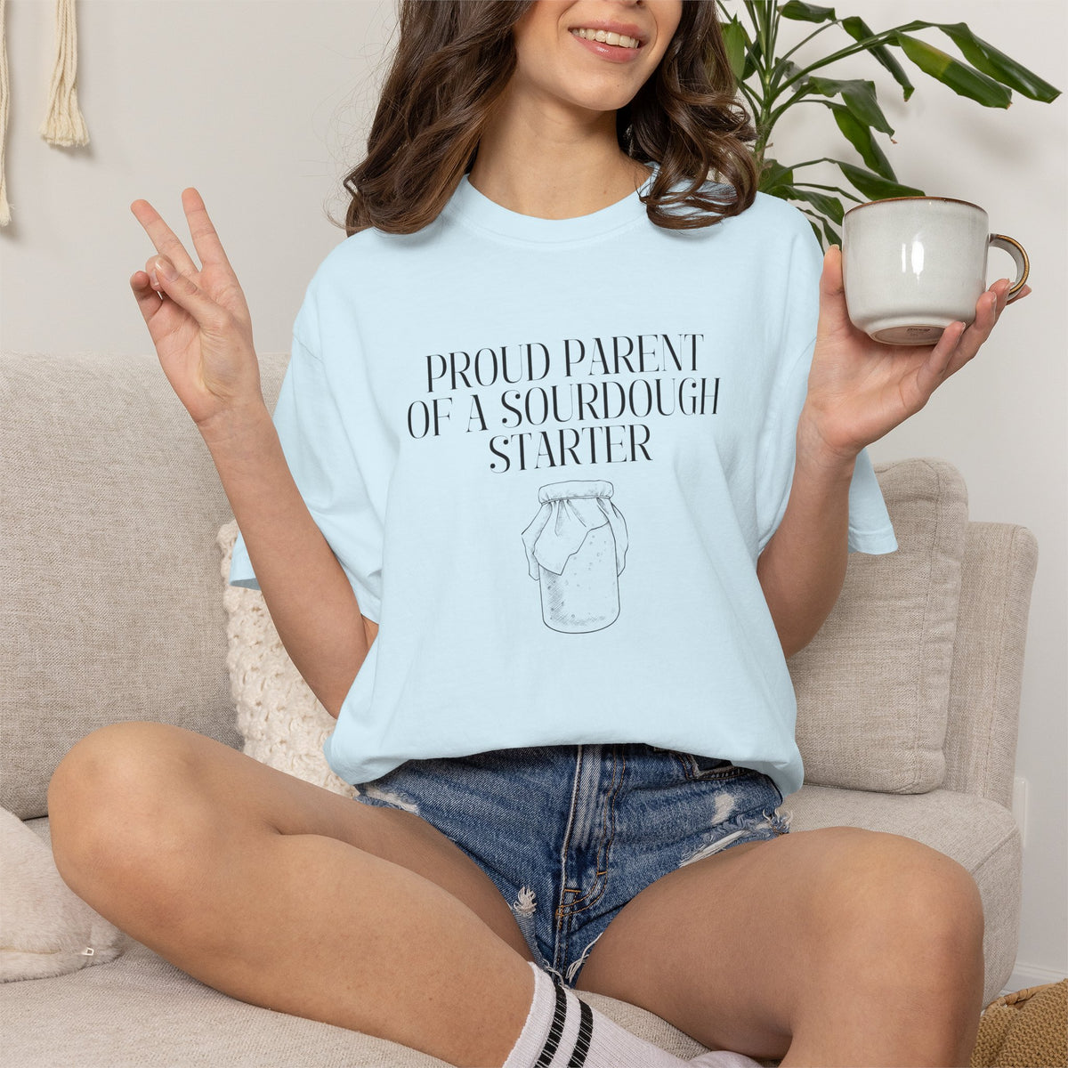 Model sitting on a couch wearing a light blue Comfort Colors® "Proud Parent of a Sourdough Starter" t-shirt with vintage sourdough jar graphic. Funny sourdough shirt for bakers, bread makers, and homemade bread enthusiasts.