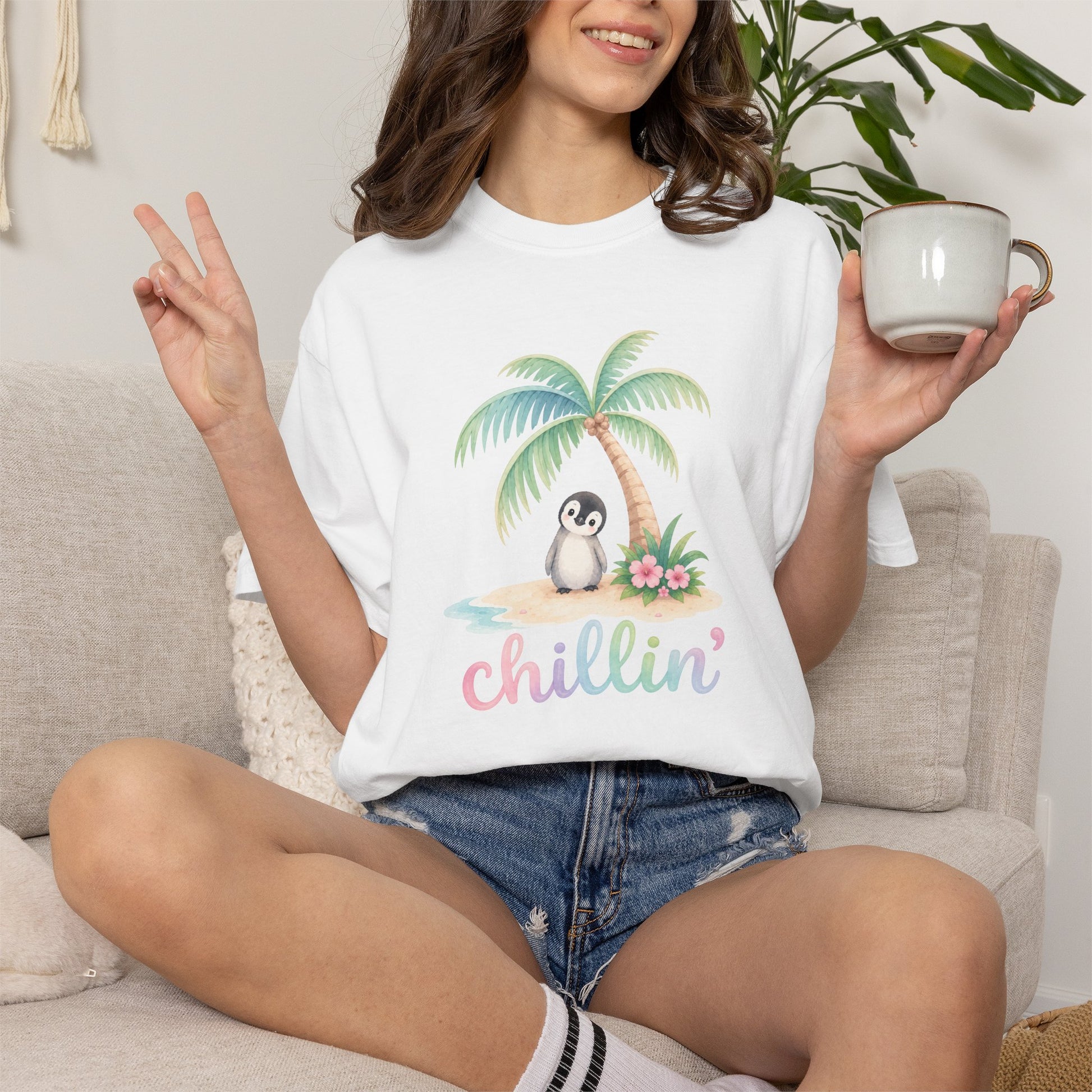 A woman sitting on a beige couch wearing a white graphic t-shirt with a cartoon penguin under a palm tree and the text 'chillin'' in pastel letters. She is holding a white mug and making a peace sign.