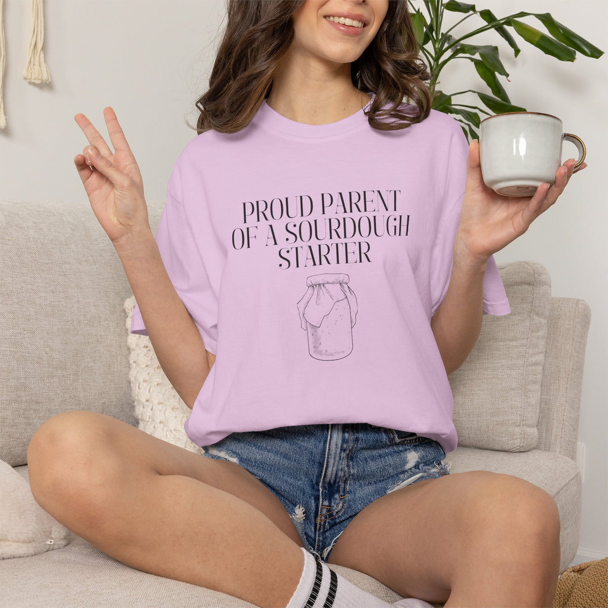 Model sitting and wearing a light pink Comfort Colors® "Proud Parent of a Sourdough Starter" t-shirt with vintage sourdough jar graphic. Funny sourdough shirt for bakers, bread makers, and homemade bread enthusiasts.