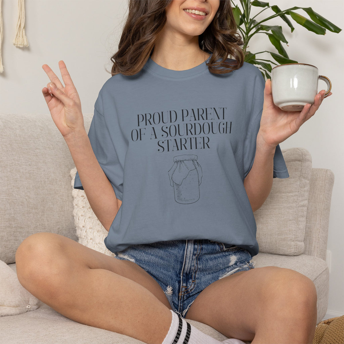 Model sitting on a couch wearing a  denim blue Comfort Colors® "Proud Parent of a Sourdough Starter" t-shirt with vintage sourdough jar graphic. Funny sourdough shirt for bakers, bread makers, and homemade bread enthusiasts.