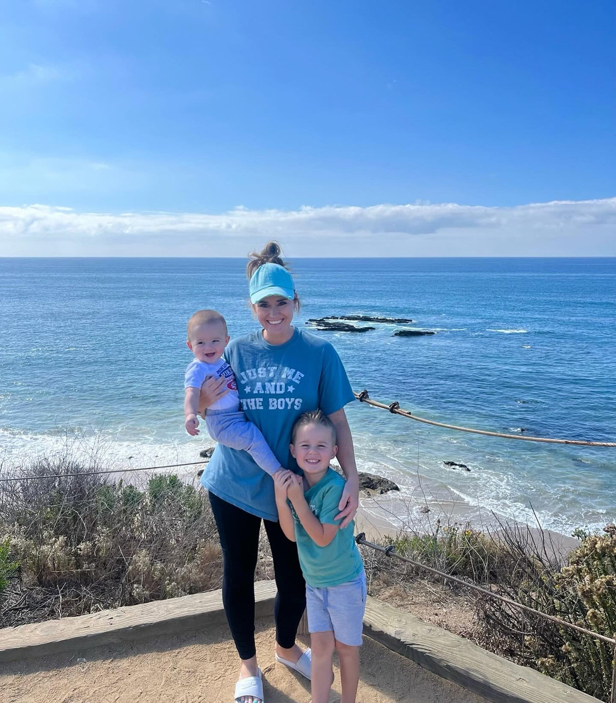 Mom of boys wearing the Just Me and the Boys tee at the beach, holding her baby with her young son beside her