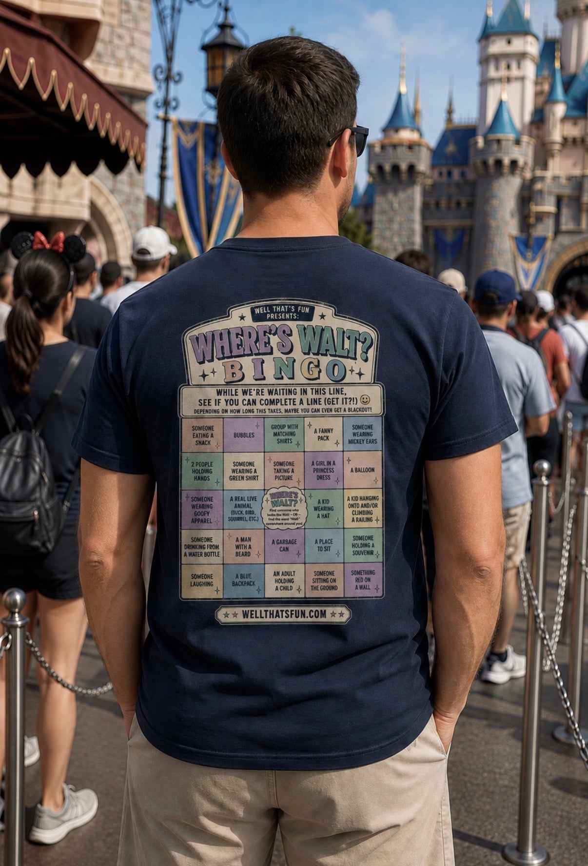 A man viewed from behind wearing a navy blue t-shirt with a large 'Where's Wait? Bingo' game chart graphic on the back, standing in a theme park queue with a castle in the background.