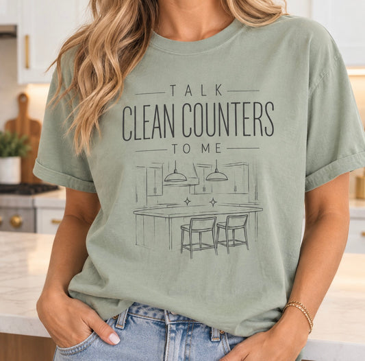 Woman in a bright kitchen wearing a sage green Talk Clean Counters To Me T-shirt with a line-drawn kitchen island and two stools