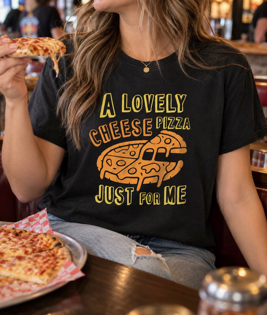 A woman eating cheese pizza while wearing a black graphic tshirt that says “a lovely cheese pizza just for me” in bright orange and yellow print with a pizza graphic 