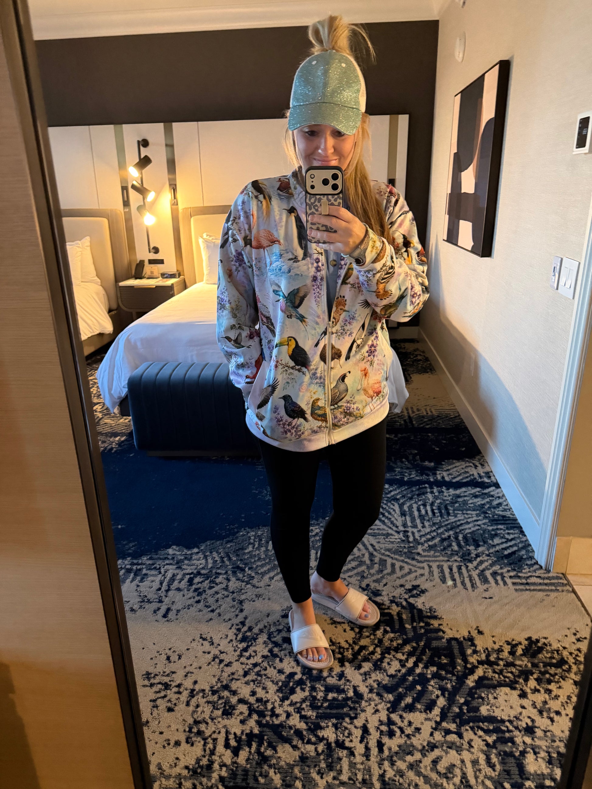 A woman takes a mirror selfie in a hotel room wearing a light blue-green baseball cap, a tie-dye zip-up jacket with a colorful bird print, black leggings, and white slide sandals.
