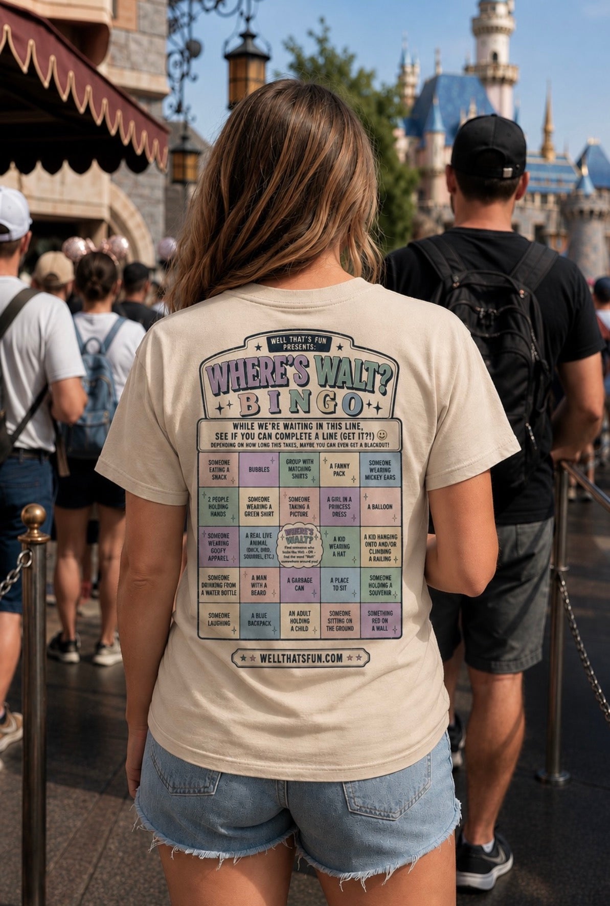 A woman seen from behind wearing a beige graphic T-shirt with a colorful 'Where's Walt? Bingo' print, standing in a theme park crowd with castles in the background.