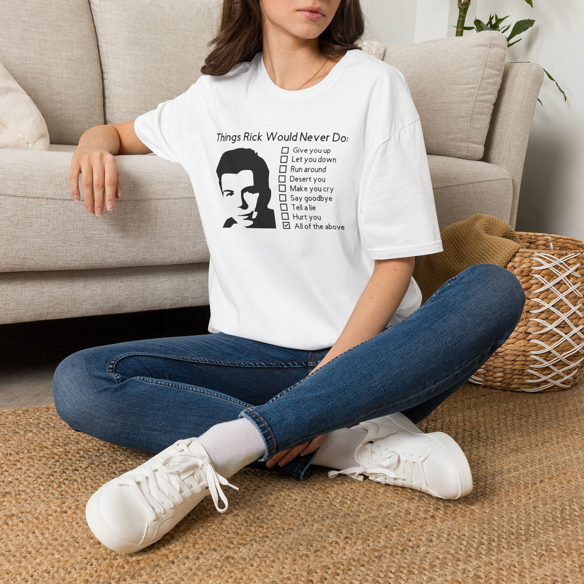 Woman seated by a couch in a white Things Rick Would Never Do t-shirt, a funny rickroll checklist tee with a Rick Astley-inspired silhouette.