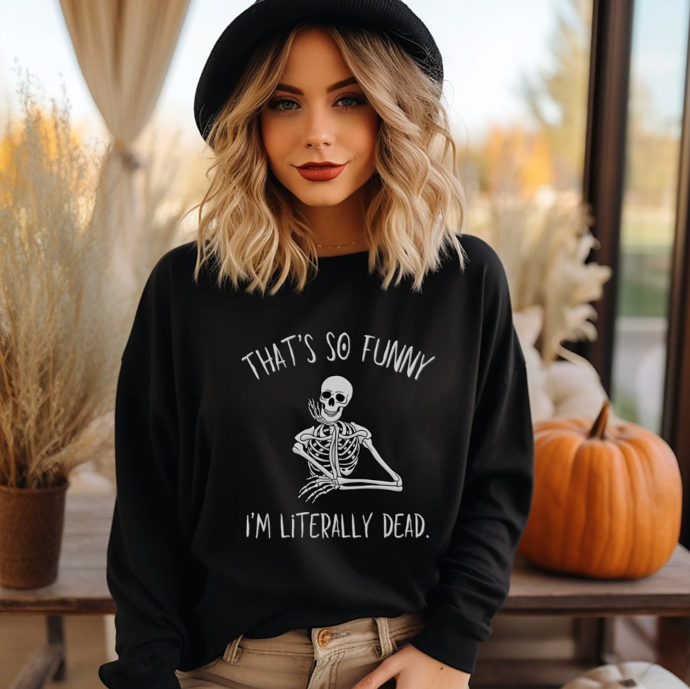 A woman wearing a black crewneck sweatshirt with a white skeleton graphic and the text 'THAT'S SO FUNNY I'M LITERALLY DEAD.' She is also wearing a black hat and tan pants, standing in a cozy, autumn-themed indoor setting with pumpkins and dried grass.