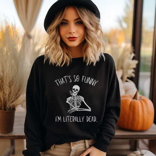 A woman wearing a black crewneck sweatshirt with a white skeleton graphic and the text 'THAT'S SO FUNNY I'M LITERALLY DEAD.' She is also wearing a black hat and tan pants, standing in a cozy, autumn-themed indoor setting with pumpkins and dried grass.