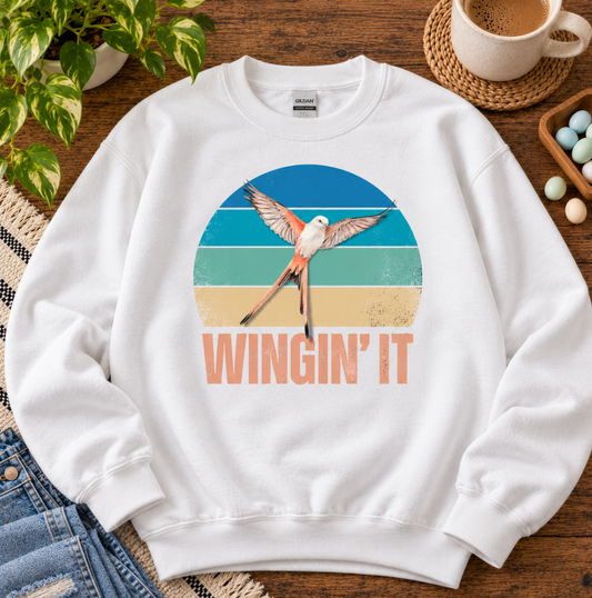 A white crewneck sweatshirt with a Wingspan-inspired scissor-tailed flycatcher and colorful retro sunset design that says "winging it"