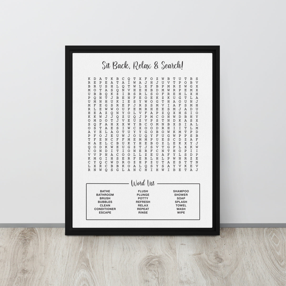 A framed bathroom themed word search canvas leaning against a light gray wall on a pale wood floor. The poster reads 'Sit Back, Relax & Search!' at the top and lists common bathroom and household words in a puzzle below.