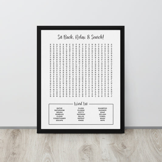 A framed bathroom themed word search canvas leaning against a light gray wall on a pale wood floor. The poster reads 'Sit Back, Relax & Search!' at the top and lists common bathroom and household words in a puzzle below.