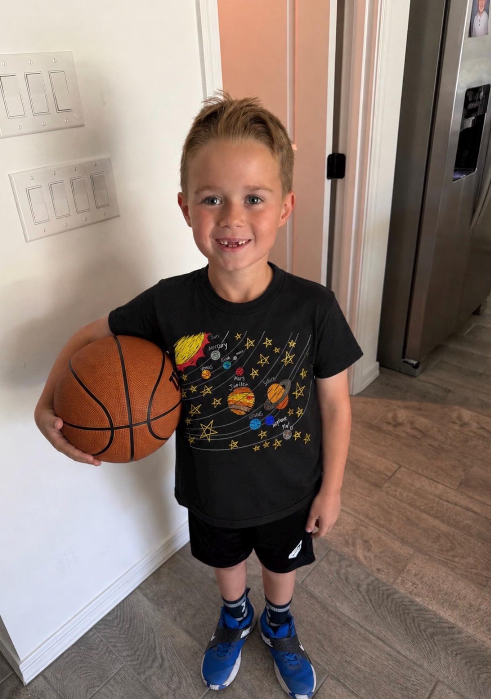 A young boy smiling and holding a basketball, wearing a black graphic t-shirt with a colorful hand drawn outer space solar system design, black shorts, and blue sneakers in a home setting.