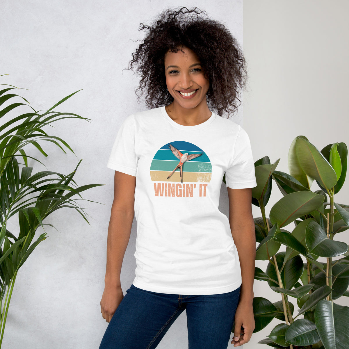 A smiling woman wearing a Wingin' It graphic t-shirt with a Wingspan-inspired scissor-tailed flycatcher over a colorful retro sunset design