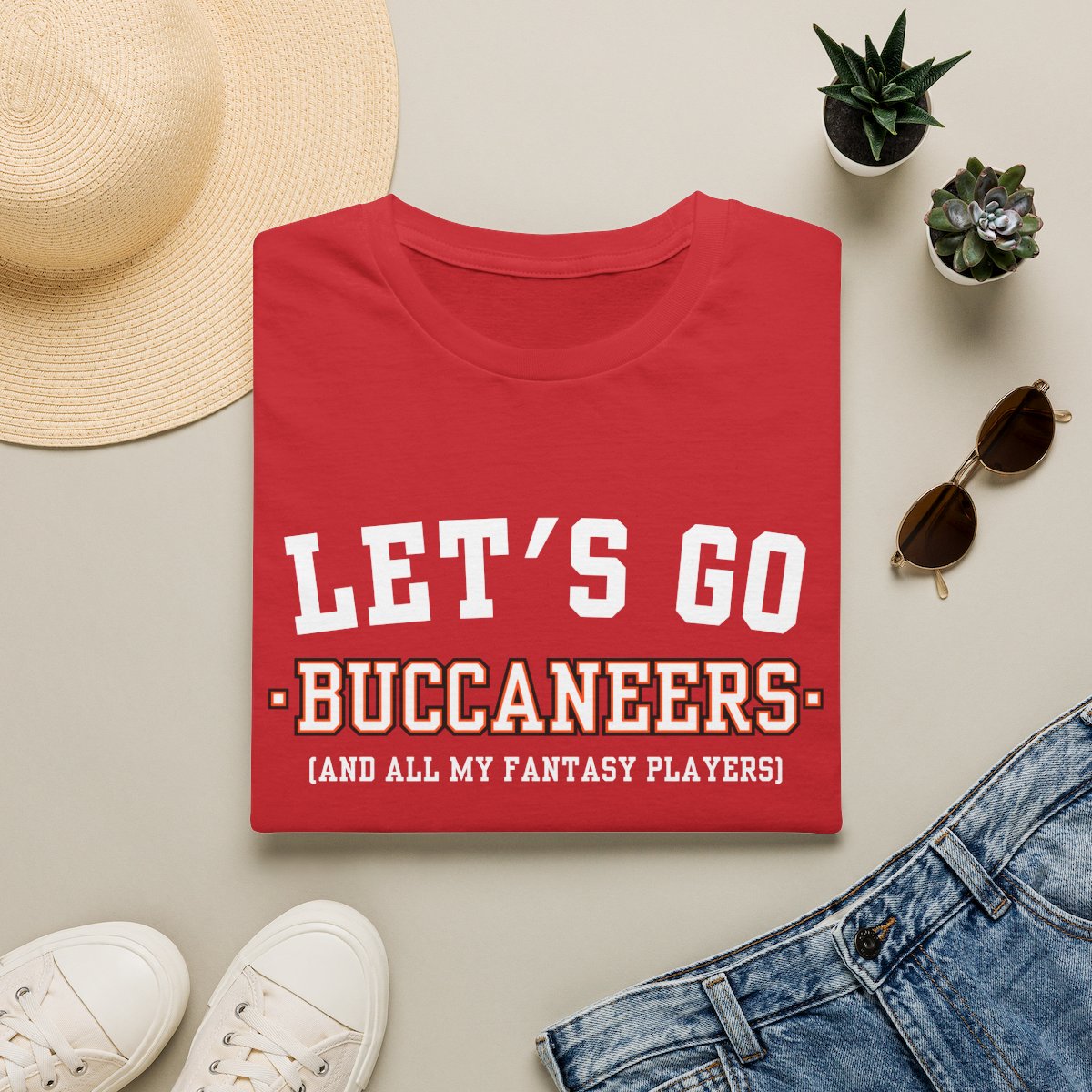 Red Buccaneers fantasy football tee folded on a neutral flat lay with a straw hat and sneakers