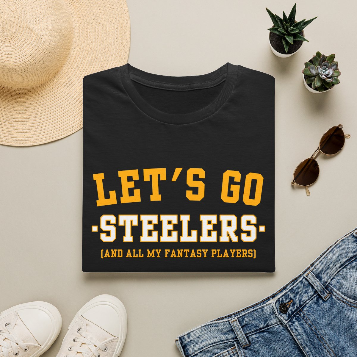 Black Steelers fantasy football tee folded on a neutral flat lay with a straw hat and sneakers