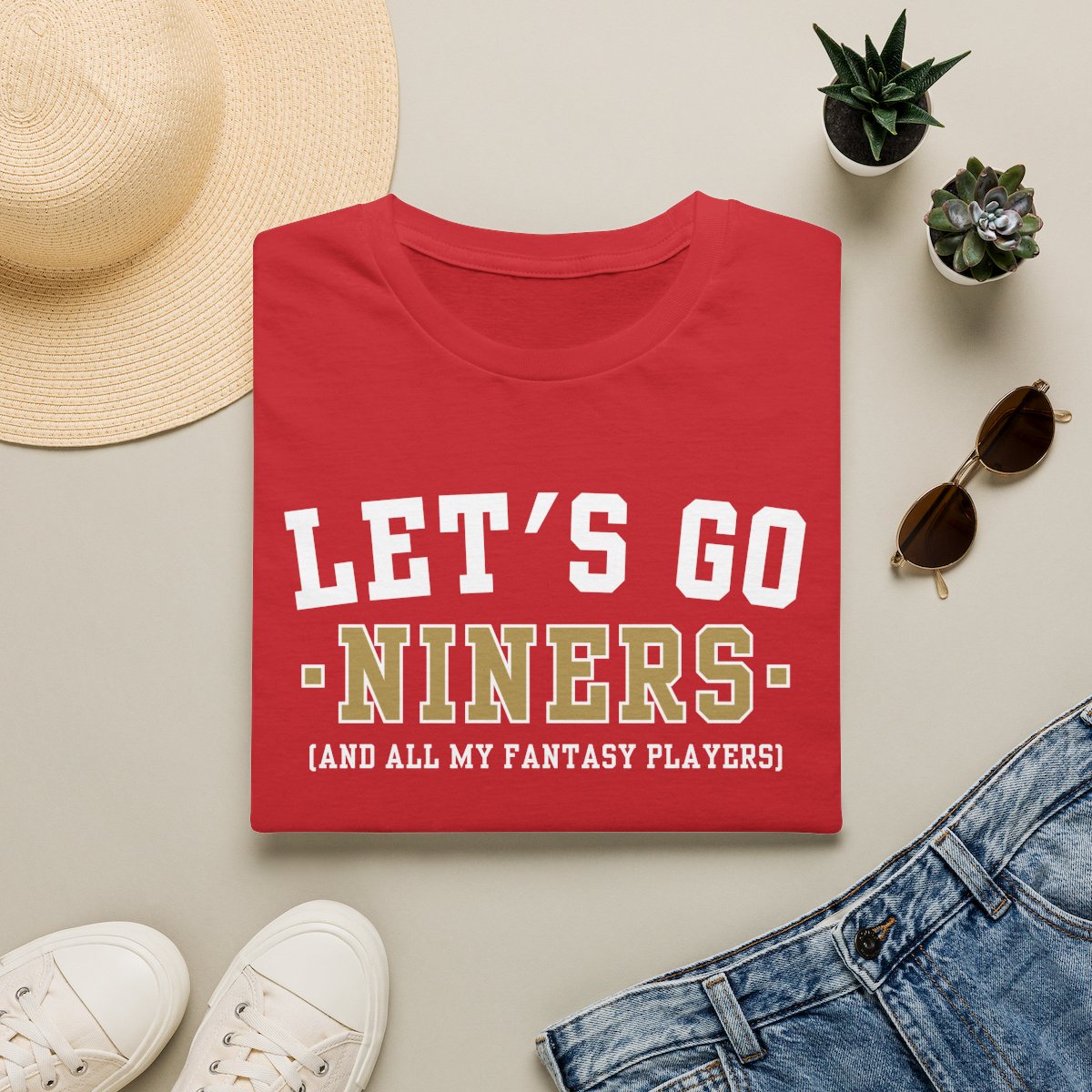 Red Niners fantasy football tee folded on a neutral flat lay with a straw hat and sneakers
