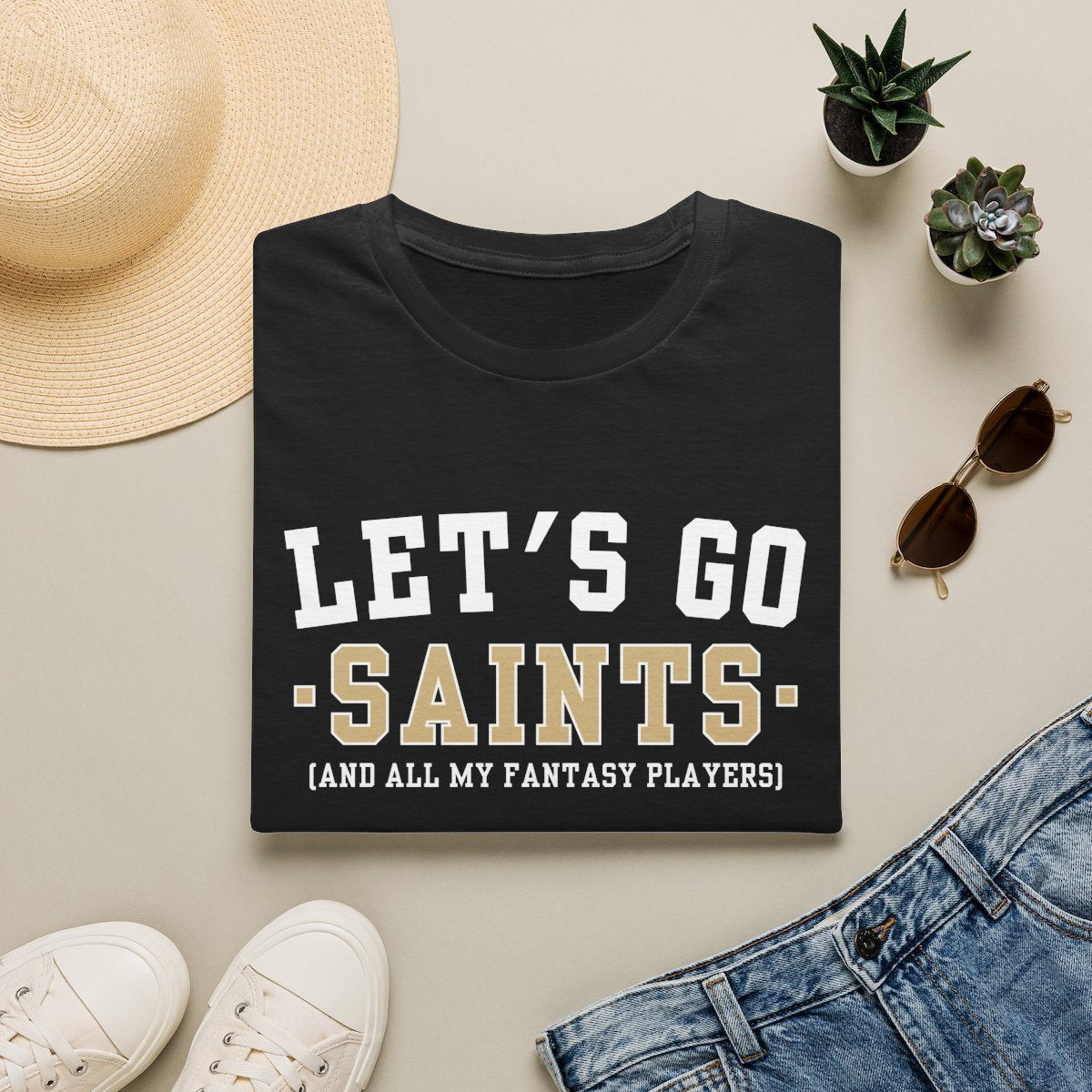 Black Saints fantasy football tee folded on a neutral flat lay with a straw hat and sneakers